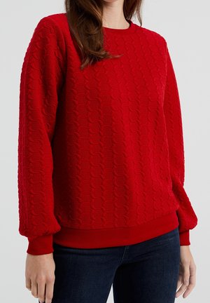 Kvinde iført en rød, tekstureret, langærmet sweater og mørkeblå jeans, stående mod en ensfarvet lys baggrund.