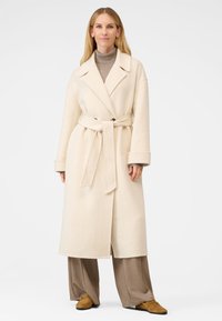 Cremefarvet uld trenchcoat med bælte i taljen, brede klapper og store frontlommer, parret med brune bukser og ruskinds fodtøj.