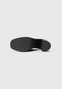 Schwarze Schuhsohle mit einer strukturierten Oberfläche, leicht erhöhtem Absatz und einer glatten, abgerundeten Zehe. Das Design ist minimalistisch und schlank.