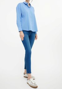 Vaaleansininen raidallinen painettava paita, rento malli, yhdistetty tummansinisiin kireisiin farkkuihin ja harmaisiin lenkkareihin, joissa on valkoiset yksityiskohdat ja ruskeat pohjat.