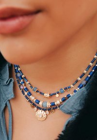 Collier multi-brins avec des perles bleues et des accents dorés, incluant un charme en forme d'œil et divers éléments décoratifs. Des surfaces texturées mettent en valeur les détails.