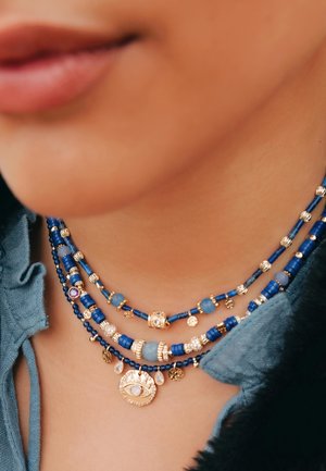 Collier multi-brins avec des perles bleues et des accents dorés, incluant un charme en forme d'œil et divers éléments décoratifs. Des surfaces texturées mettent en valeur les détails.