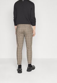 Persoon in slim-fit beige geruite broek, zwarte sokken, zwarte leren schoenen en een zwart longsleeve shirt, staande tegen een effen achtergrond.