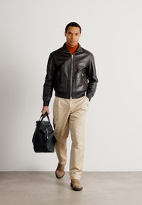 Giacca bomber nera in pelle con polsini a coste, dolcevita arancione, pantaloni beige e stivali alla caviglia marroni. Porta una borsa da viaggio nera.