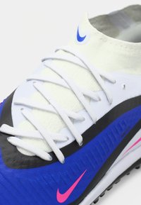 Αθλητικό παπούτσι με λευκό και μπλε επάνω μέρος, μαύρες λεπτομέρειες και ροζ διακοσμήσεις. Διαθέτει υλικό με υφή και λευκά κορδόνια.