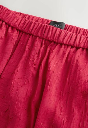 Red, textured fabric with a gathered elastic waistband. Tag displays the brand name "NEXT" in a contrasting color.