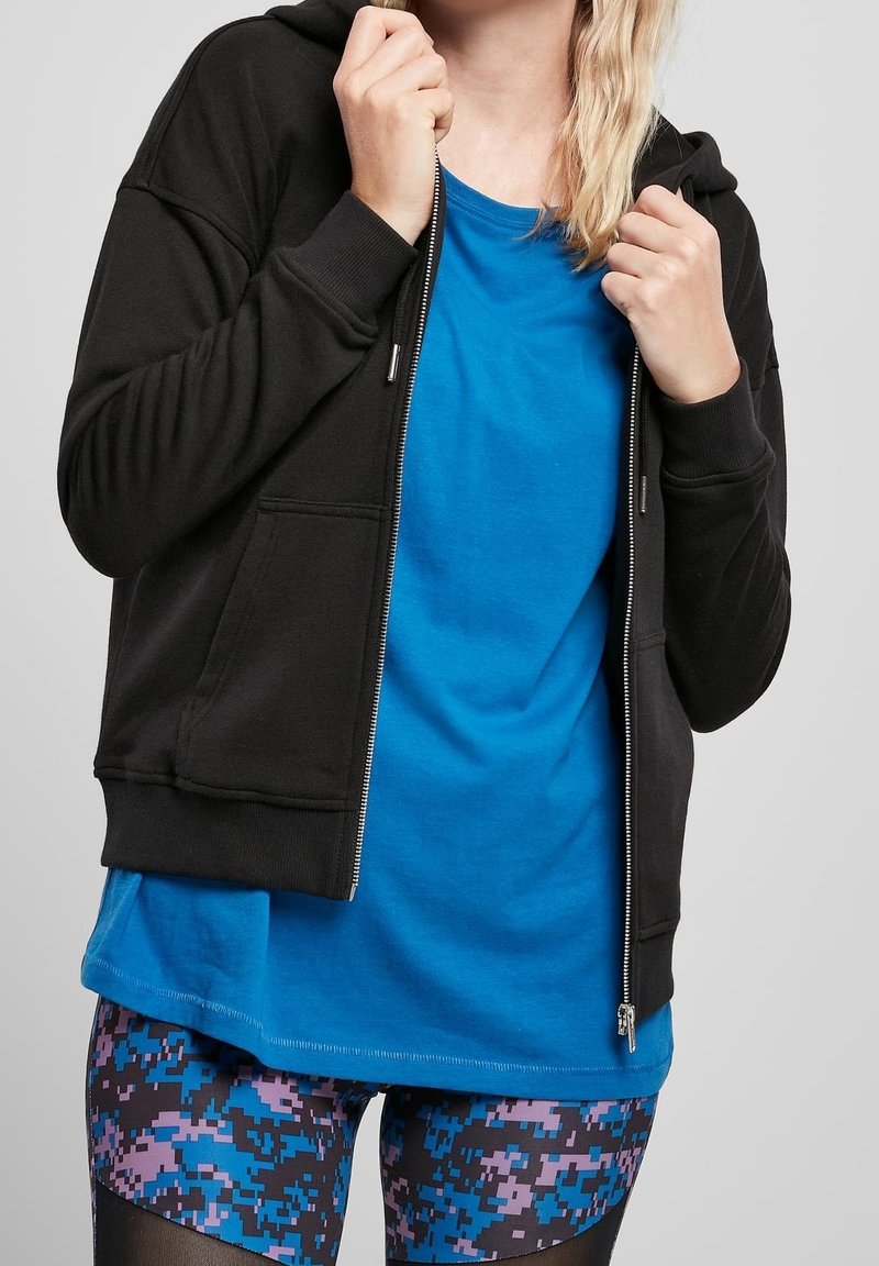 Persona che indossa un hoodie nera con zip sopra una maglietta blu e leggings pixelati nei colori blu, viola e nero, mentre tiene il collo dell'hoodie.