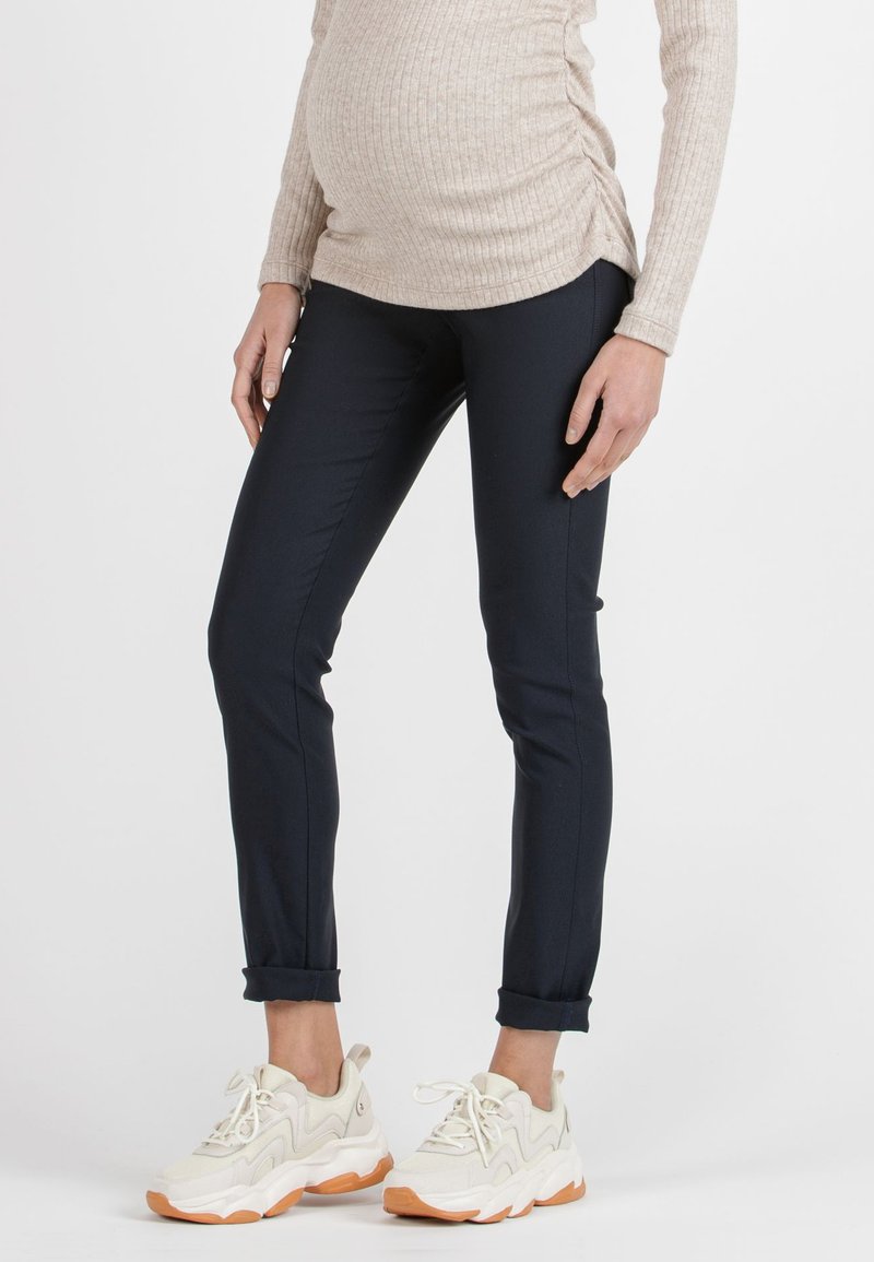 Navy maternity pants with a slim fit, soft stretch fabric, and rolled cuffs, paired with chunky beige sneakers and a ribbed beige top.