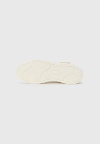 Semelle de chaussure de sport couleur crème avec une surface en caoutchouc texturée, ornée de petits motifs circulaires et d'un élément rectangulaire central pour une meilleure adhérence.