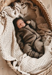 Brauner Wollfleece-Einteiler und passender Hut an einem Baby, das in einem geflochtenen Korb liegt, ausgelegt mit einer hellbeigen, gestrickten Decke.