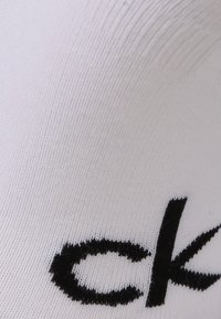 Biela tkanina z bavlny s prídavkom iných vlákien má textúrovaný vzor s čierne vyšívaným logom "ck", pričom sa vyznačuje hladkým a mäkkým povrchom.