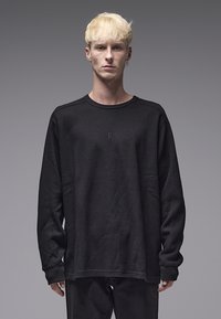 Camisa preta de manga comprida com padrão texturizado em malha tipo waffle. Apresenta decote redondo e corte solto. Exibida sobre um fundo neutro.