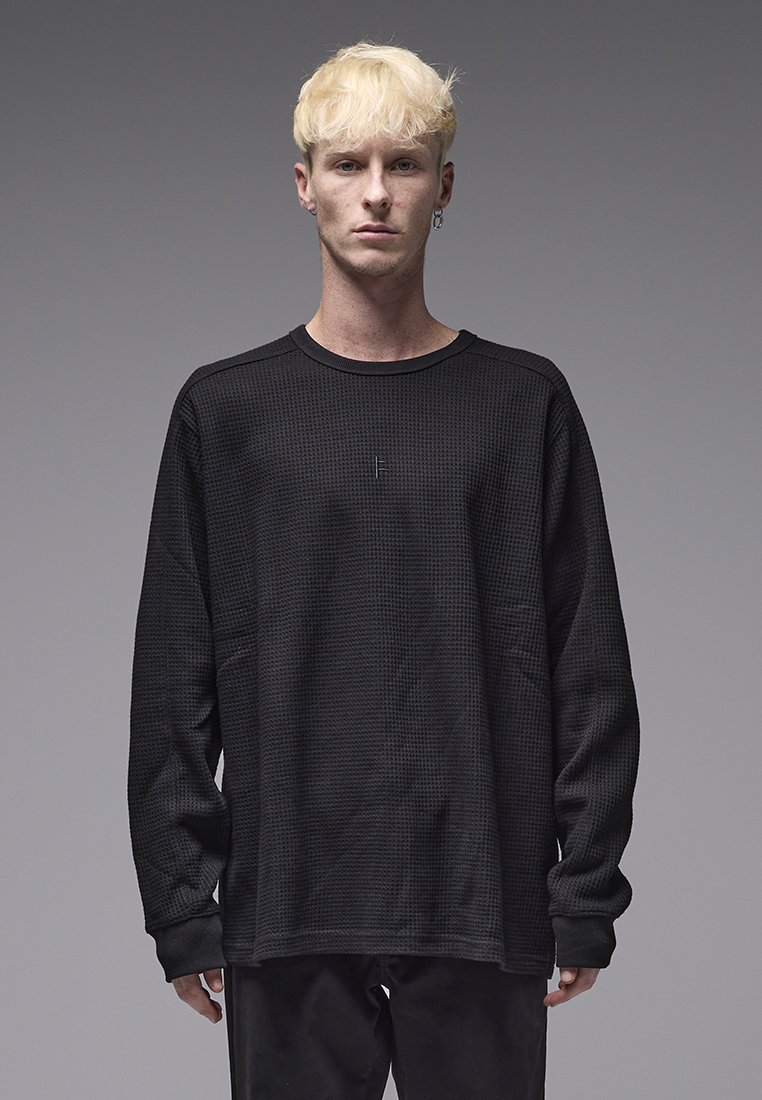 Camisa preta de manga comprida com padrão texturizado em malha tipo waffle. Apresenta decote redondo e corte solto. Exibida sobre um fundo neutro.