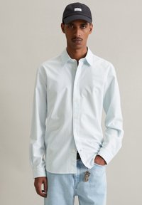 Lichtblauw gestreept overhemd met puntige kraag, gecombineerd met lichtgekleurde denim jeans en een donkere pet, met een sleutelhanger zichtbaar bij de taille.