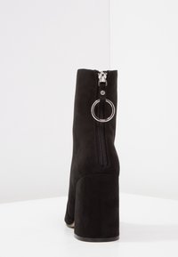Bottines en suède noir avec un talon épais, dotées d'une fermeture éclair à l'arrière et d'un accent de tirette de fermeture éclair circulaire. Texture lisse, design ajusté.