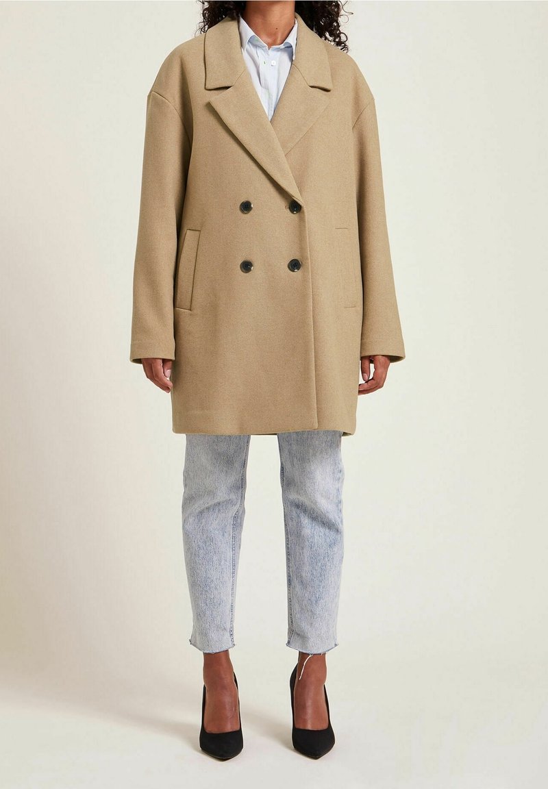 Femme portant un manteau camel oversize à double boutonnage, une chemise bleu clair, un jean délavé clair et des talons noirs à bout pointu, debout sur un fond uni.