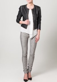 Giacca bomber in pelle nera con borchie argentate, abbinata a una camicetta bianca asimmetrica e pantaloni skinny grigi. Gli accessori includono tacchi neri.