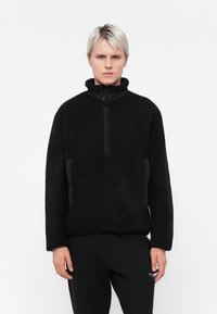 Black fleece pullover with a high collar and half-zip front. Features textured fabric, side pockets with black accent panels, and relaxed fit.