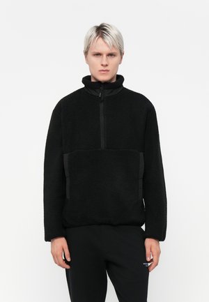 Black fleece pullover with a high collar and half-zip front. Features textured fabric, side pockets with black accent panels, and relaxed fit.