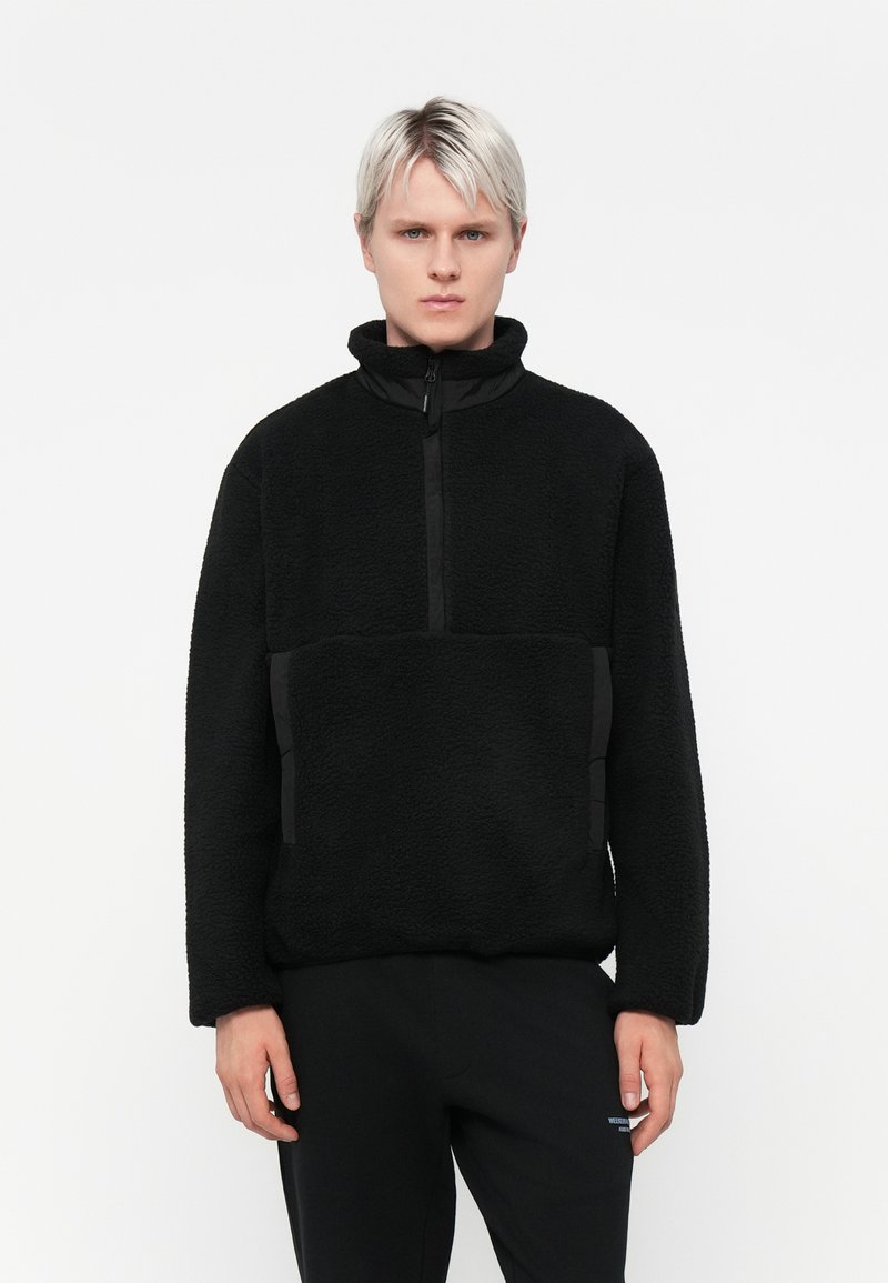 Black fleece pullover with a high collar and half-zip front. Features textured fabric, side pockets with black accent panels, and relaxed fit.