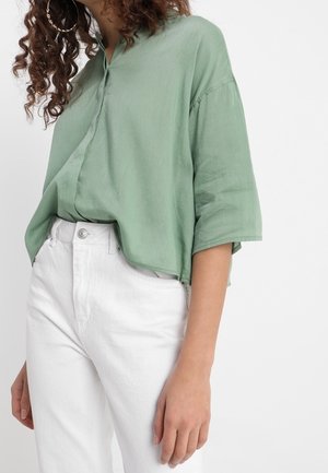 Woman wearing oversized light green button-up shirt with elbow-length sleeves and high-waisted white pants, standing against white background.