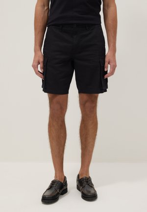 Mann trägt schwarze Cargoshorts und schwarze Leder-Schnürschuhe, steht vor schlichtem hellem Hintergrund.
