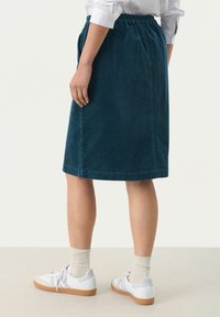 Blaue Cordrock mit elasticem Bund, knielang und subtilen vertikalen Rippungen. Kombiniert mit weißen Turnschuhen und hellen Socken.