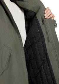 Chaqueta verde oliva con cierre de cremallera, que presenta un forro interior negro acolchado con patrones geométricos y un bolsillo lateral.