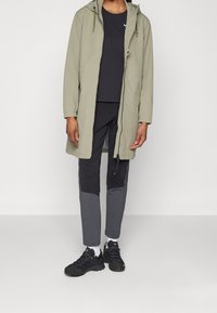 Světle zelený dlouhý kabát s kapucí, vrstvený přes černé tričko, v kombinaci s dvoubarevnými šedými kalhotami a černými sportovními botami.