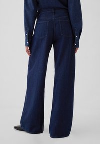 Magas derekú, széles szárú sötétkék farmernadrág, két hátsó zsebbel és finom varrási részletekkel. Simaságot és masszív textúrát kínál.