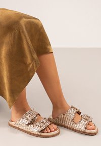 Sandalias deslizantes tejidas en dorado metálico con doble hebilla, suelas planas y una superficie texturizada. Combinadas con una falda de tono dorado.