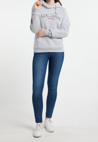Grijze hoodie met "DREIMASTER" in zwart en rood gedrukt, gecombineerd met donkerblauwe skinny jeans en witte sneakers, staand op een lichte achtergrond.