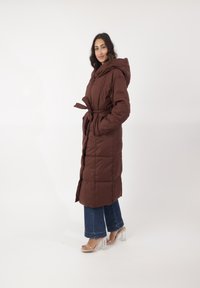 Bruine pufferjas met een capuchon, ceintuur in de taille en zijzakken. Kenmerken zijn het doorstikte ontwerp en lange mouwen, gedragen over een blauwe wijde jeans en doorzichtige hakken.