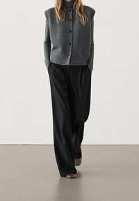 Harmaa neuleliivi napilla ja ribatulla helmalla, yhdistettynä mustiin raidallisiin leveälahkeisiin housuihin. Sileä pinta, rennon mallinen.