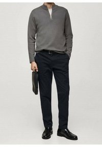 Grauer Herrenpullover mit halbem Reißverschluss am Kragen, kombiniert mit dunkelblauen Navy-Hosen. Schwarze, polierte Schuhe und eine schwarze Clutch. Glatte Stoffstruktur.