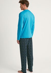 Langärmliges türkisfarbenes Oberteil kombiniert mit navyblauen Pyjamahosen, die ein grünes Blattmuster aufweisen. Lockere Passform, weicher Stoff und leichtes Material.