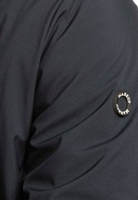 Tessuto della giacca leggero e nero con una texture liscia. Presenta una toppa rotonda nera con il testo "MARKUP" sulla manica.