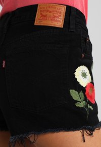 Pantalones cortos de mezclilla negra con dobladillos deshilachados, con bordado floral colorido en un lado y una etiqueta de marca de cuero marrón en la parte trasera.