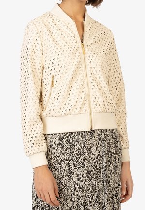 Veste bomber beige avec motif découpé triangulaire, détails de fermeture éclair dorée, portée sur une jupe à motif noir et blanc.