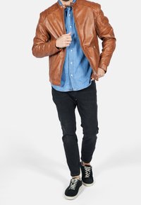 Braune Lederjacke mit gesteppten Schultern, getragen über einem hellblauen Denimhemd. Kombiniert mit schwarzen Jeans und schwarzen Sneakers mit weißen Sohlen.