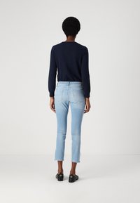 Persona con cabello corto y rizado, vistiendo un suéter azul marino, jeans cortos azul claro y mocasines negros, de pie frente a un fondo blanco liso.