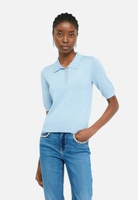 Polo shirt bleu clair à manches courtes, en tissu texturé, avec trois boutons dorés et un petit logo sur la poitrine gauche. Associé à un jean.