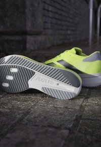 Lysegule Adidas løbesko med "Adizero" såldetalje hvilende på stensat belægning mod en mørk, tekstureret væg.