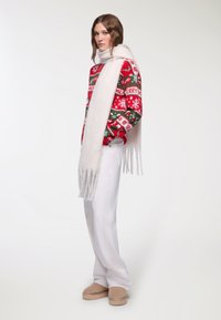 Czerwony i zielony sweter w wzory z płatkami śniegu, biały szalik, białe spodnie i beżowe buty wsuwane. Szalik ma frędzlaste końce.