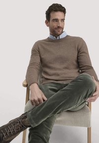 Beige gebreide trui, lichtblauw overhemd met kraag eronder, groene corduroy broek, bruine veterschoenen, zittend op een lichtgekleurde stoel met textuur.