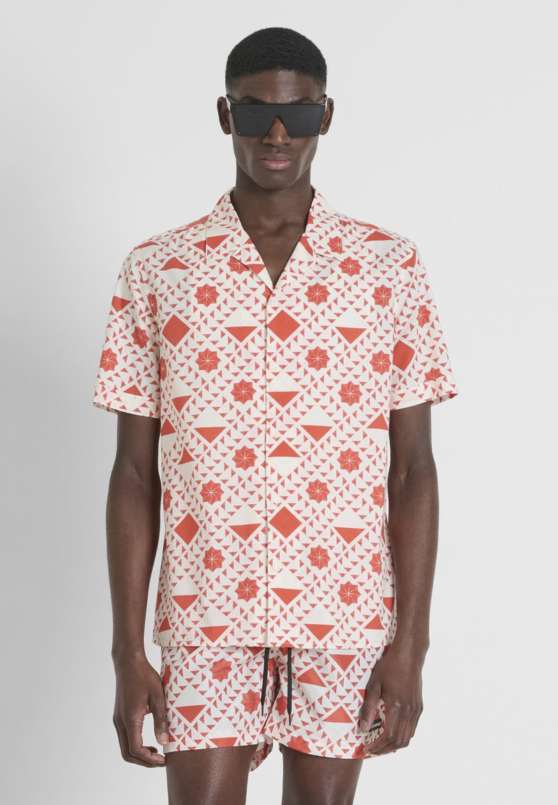 Korte mouwen shirt met een geometrisch rood-wit patroon, gecombineerd met bijpassende shorts, gemaakt van lichtgewicht stof. Draagt een zonnebril met een donker montuur.