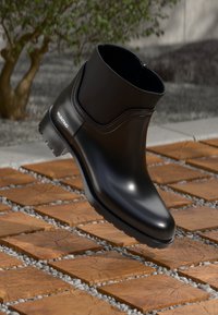 Bota de tornozelo impermeável preta com um acabamento suave, bico arredondado e sola texturizada. Apresenta um detalhe de logotipo discreto e um design minimalista.