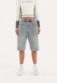 Shorts de mezclilla en azul claro, hasta la rodilla, con un corte holgado, bolsillos frontales y un sutil detalle de logo bordado. Combinados con zapatillas blancas.