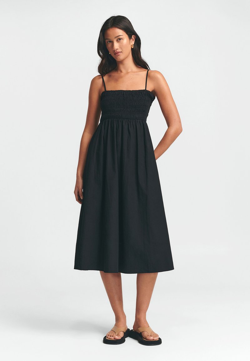 Vestido negro con un corsé fruncido, tirantes finos y una falda fluida en forma de A. La tela es ligera, adecuada para climas cálidos.