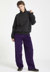 Sort pullover-hoodie med knappedetaljer og snoretræk. Kombineret med vidbenede lilla kordfløjlsbukser med ribbet tekstur.